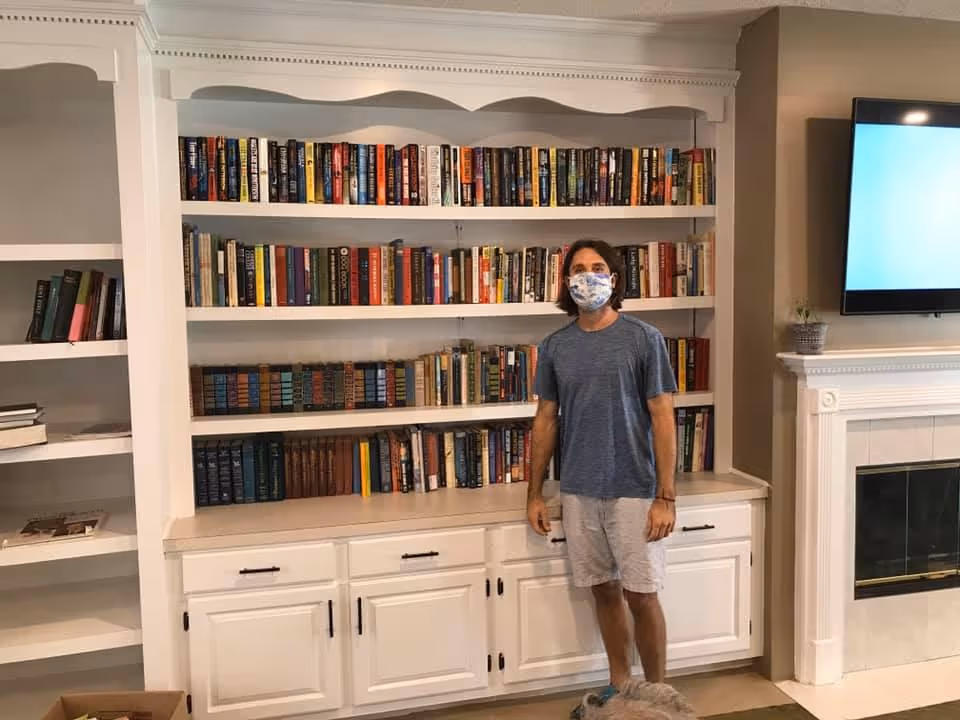 A man wearing a blue t-shirt, light-colored shorts, and a face mask stands in front of a large white bookshelf filled with books. To the right, there is a white fireplace with a small plant on the mantel and a flat-screen TV mounted above it.