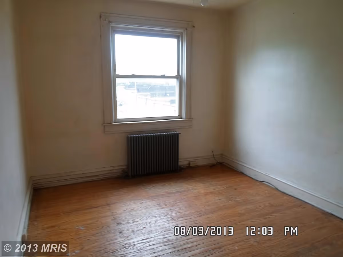 Empty room with wooden floor, plain white walls, a single window with white trim, and a black radiator beneath the window.