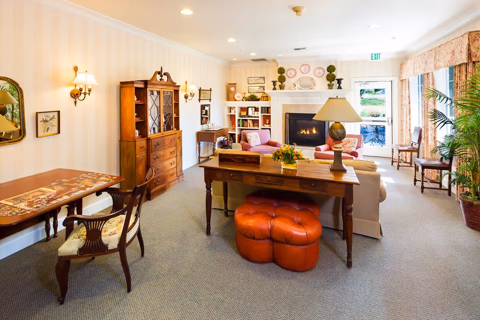 A cozy living room area in a memory care community featuring a fireplace with decorative plates and plants on the mantel. The room has comfortable seating including two pink armchairs and a beige sofa. There is a wooden desk with a lamp and a red cushioned ottoman underneath. To the left, there is a wooden cabinet and a table with a partially completed puzzle and a chair. Large windows with floral curtains allow natural light to brighten the room, and there are several plants adding greenery.