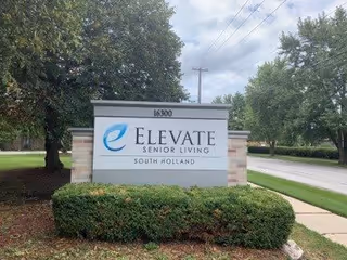 Outdoor view of a sign for Elevate Senior Living South Holland, located at 16300. The sign is surrounded by green bushes and trees, with a sidewalk and road visible in the background under a cloudy sky.