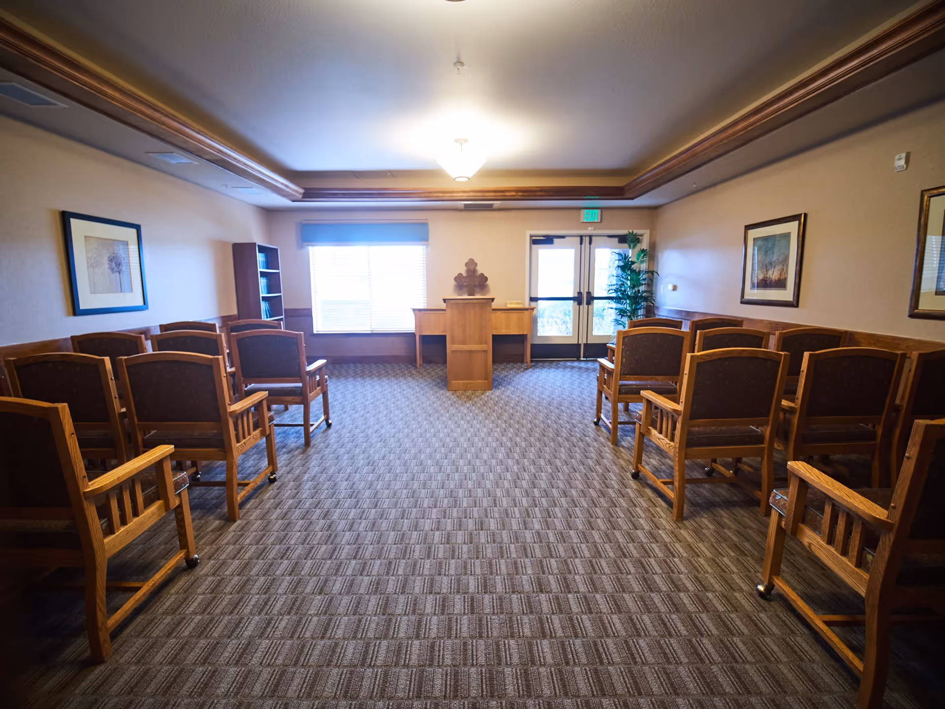 A small chapel or meeting room with rows of wooden chairs facing a wooden podium. The room has beige walls, carpeted floor, framed artwork on the walls, a window with blinds, and a double glass door with an exit sign above it. There is also a bookshelf and a potted plant near the door.