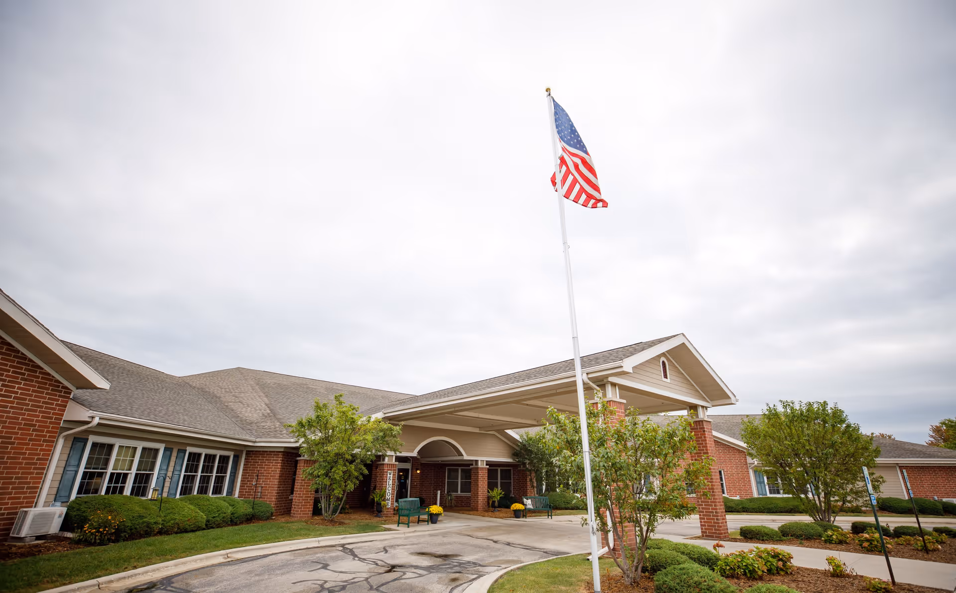Exterior view of a senior living facility building with a covered entrance, an American flag on a flagpole, and landscaped greenery including bushes and small trees under a cloudy sky.