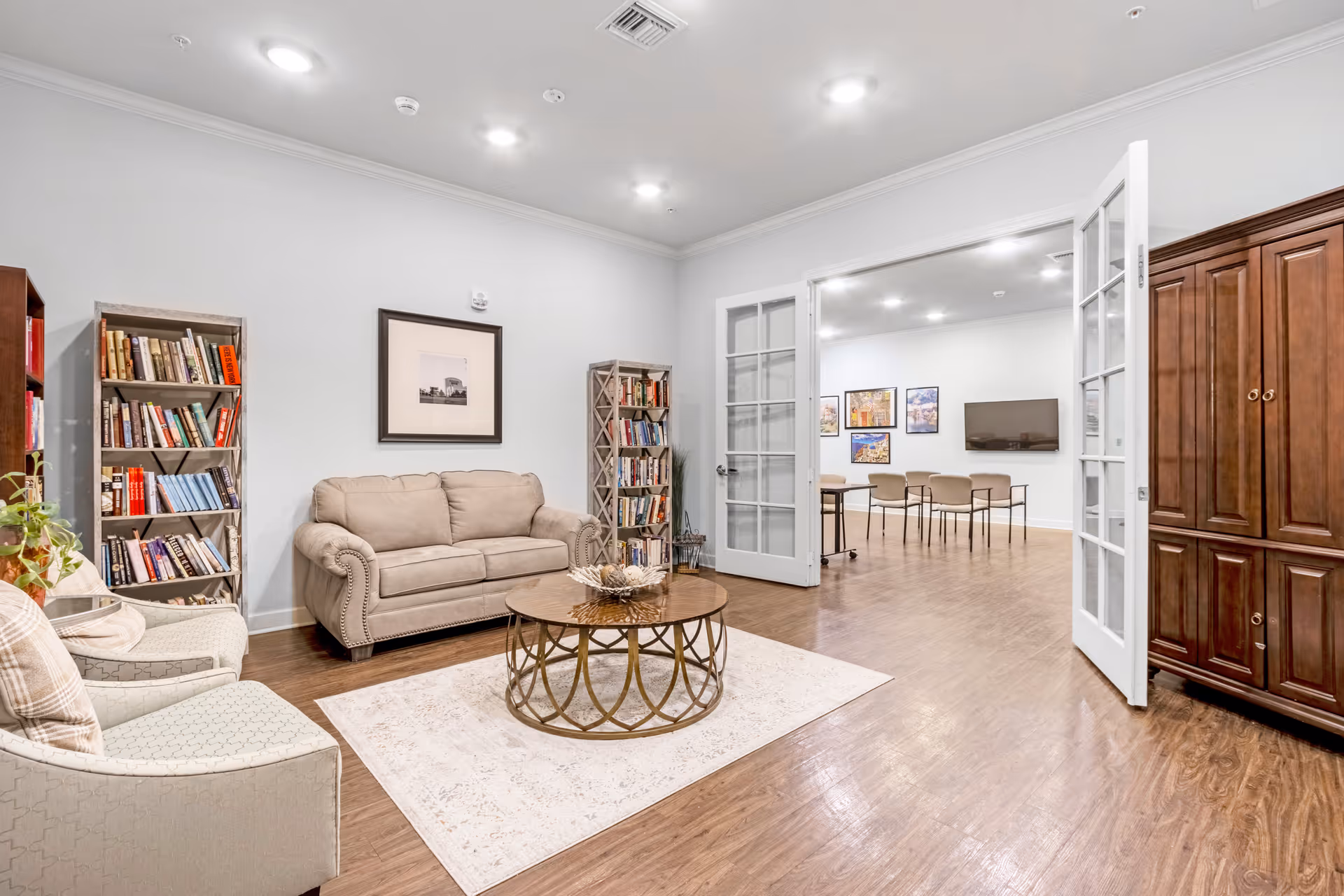 A cozy living room area with a beige couch, two armchairs, a round coffee table on a light rug, and two tall bookshelves filled with books. The room has wooden flooring and white walls with a framed picture above the couch. French doors open to an adjacent room with tables, chairs, framed pictures on the wall, and a wall-mounted TV.