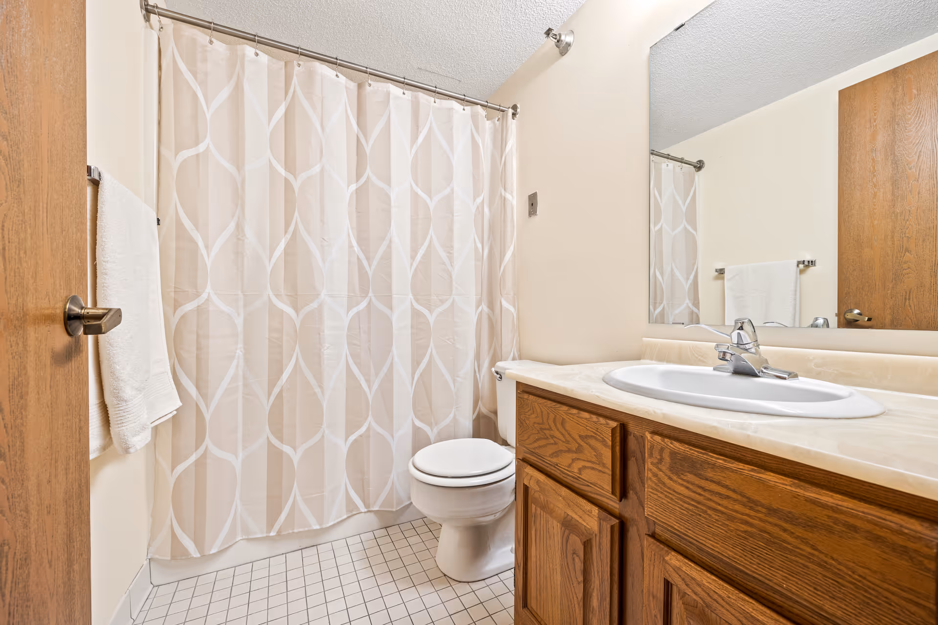 A clean bathroom with a white toilet, a wooden vanity with a sink and faucet, a large mirror above the sink, a beige and white patterned shower curtain, a towel hanging on a rack next to the door, and tiled floor.