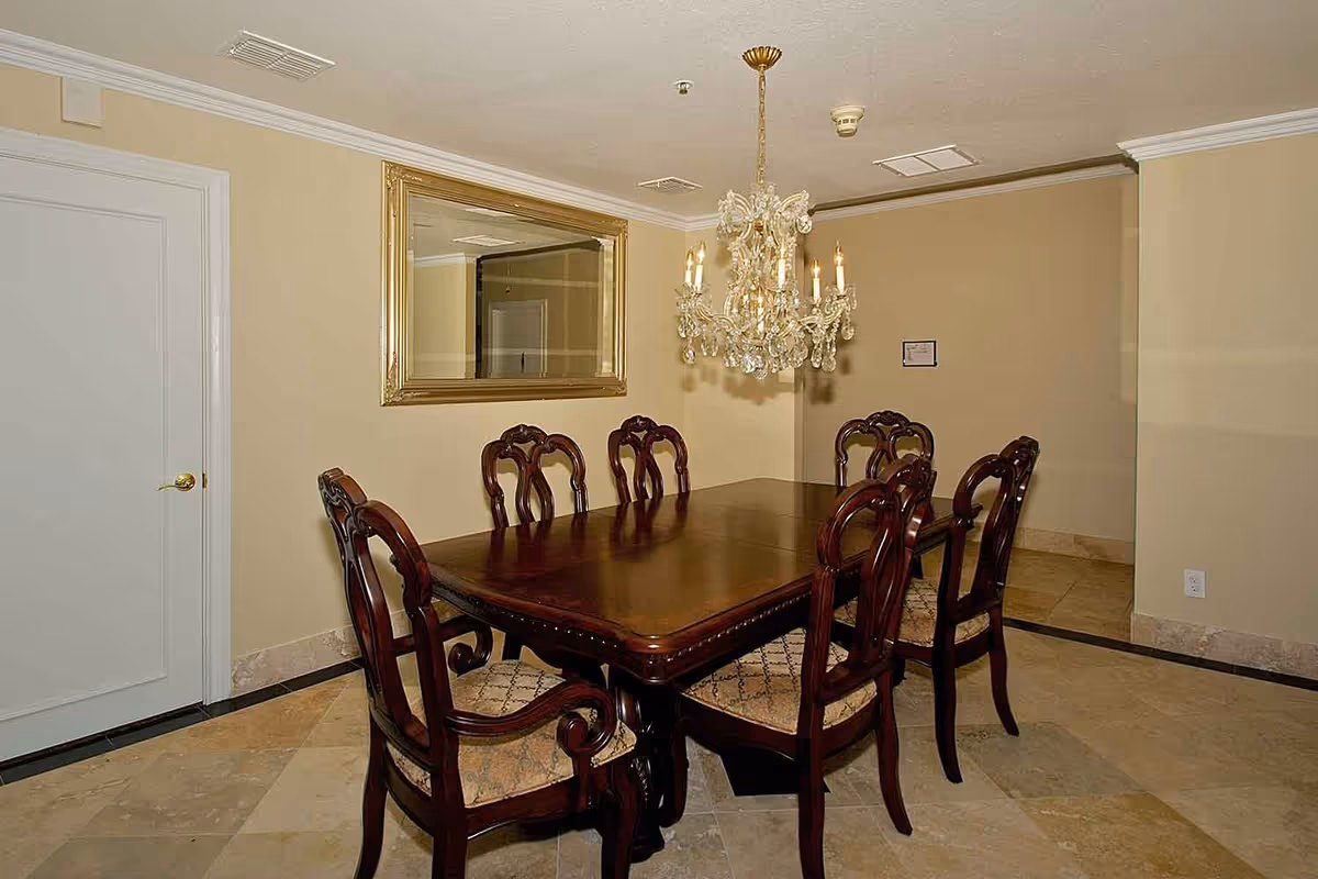 A formal dining room with a dark wooden rectangular table surrounded by six matching chairs with upholstered seats. A large ornate chandelier hangs above the table, and a large gold-framed mirror is mounted on the beige wall. The floor is tiled with a light-colored stone, and there is a white door with a gold handle on the left side of the room.