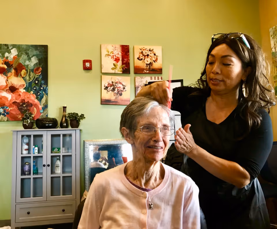 An elderly woman with glasses and a light pink cardigan is seated while a caregiver or hairstylist combs her short gray hair in a cozy room decorated with floral paintings and a cabinet with various items.