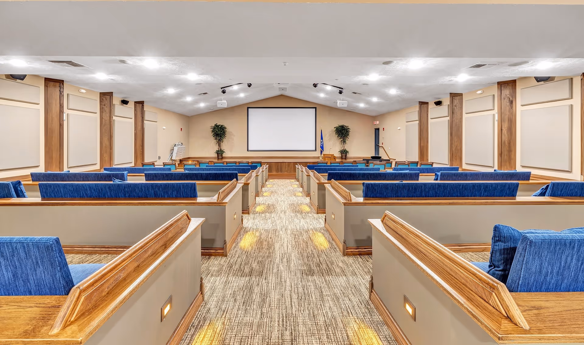 Interior view of a large meeting or presentation room with rows of wooden pew-style seating featuring blue cushions, a central aisle with carpeted flooring, and a large blank projection screen at the front flanked by two potted plants and a podium.