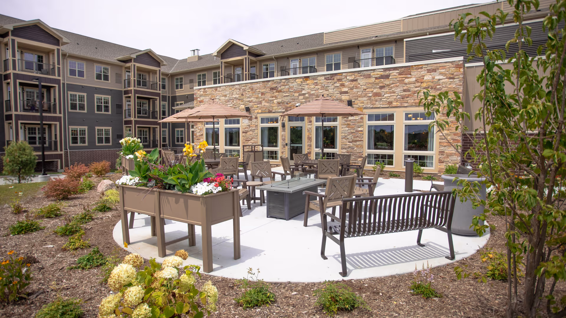 Outdoor patio area at a senior living facility with multiple chairs, tables, umbrellas, and a fire pit surrounded by landscaped garden beds with flowers and shrubs. The building in the background has stone and siding exterior with multiple windows.