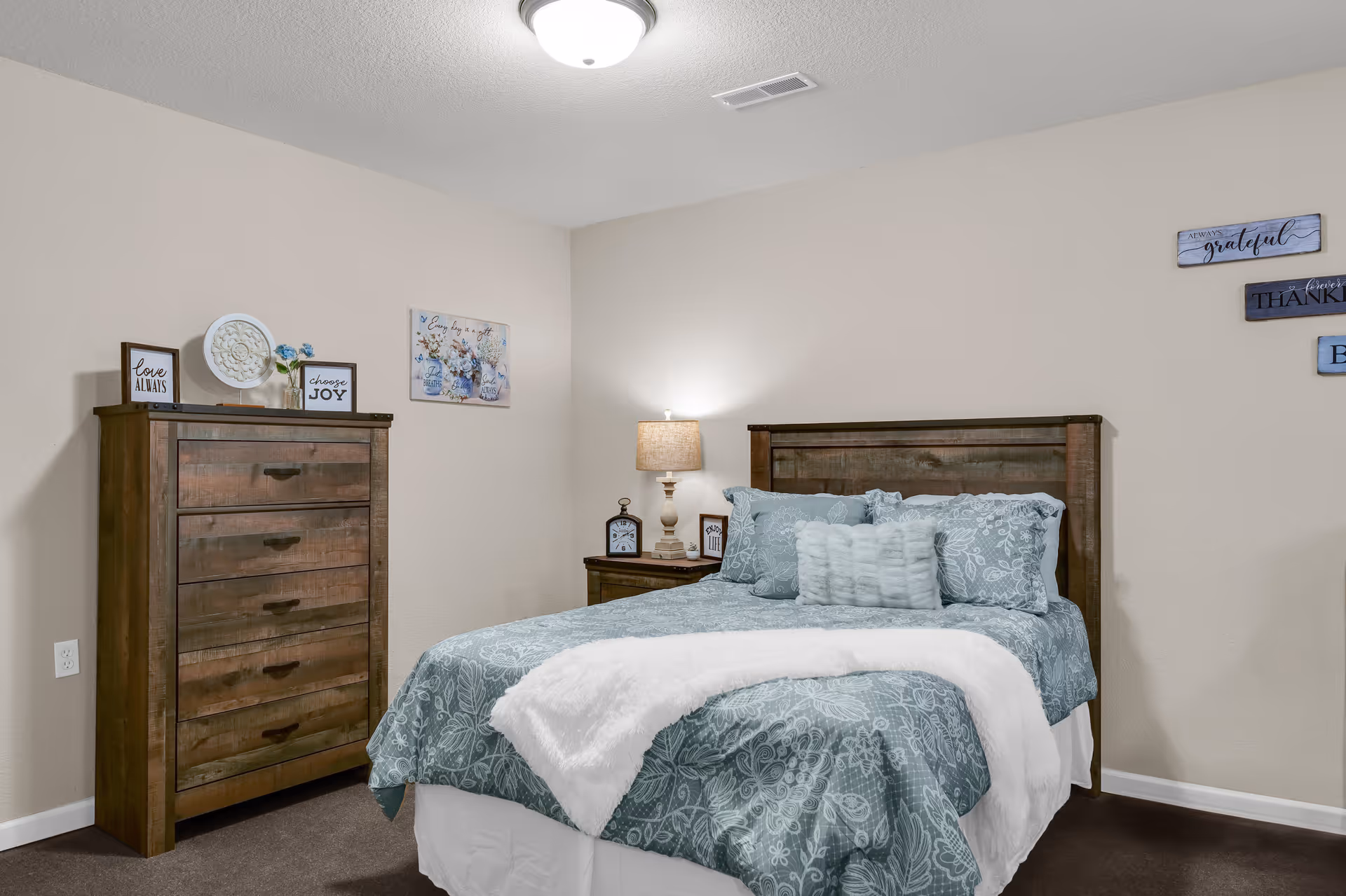Cozy bedroom with a made bed, wooden headboard and dresser, nightstand with a lamp, and wall decorations.