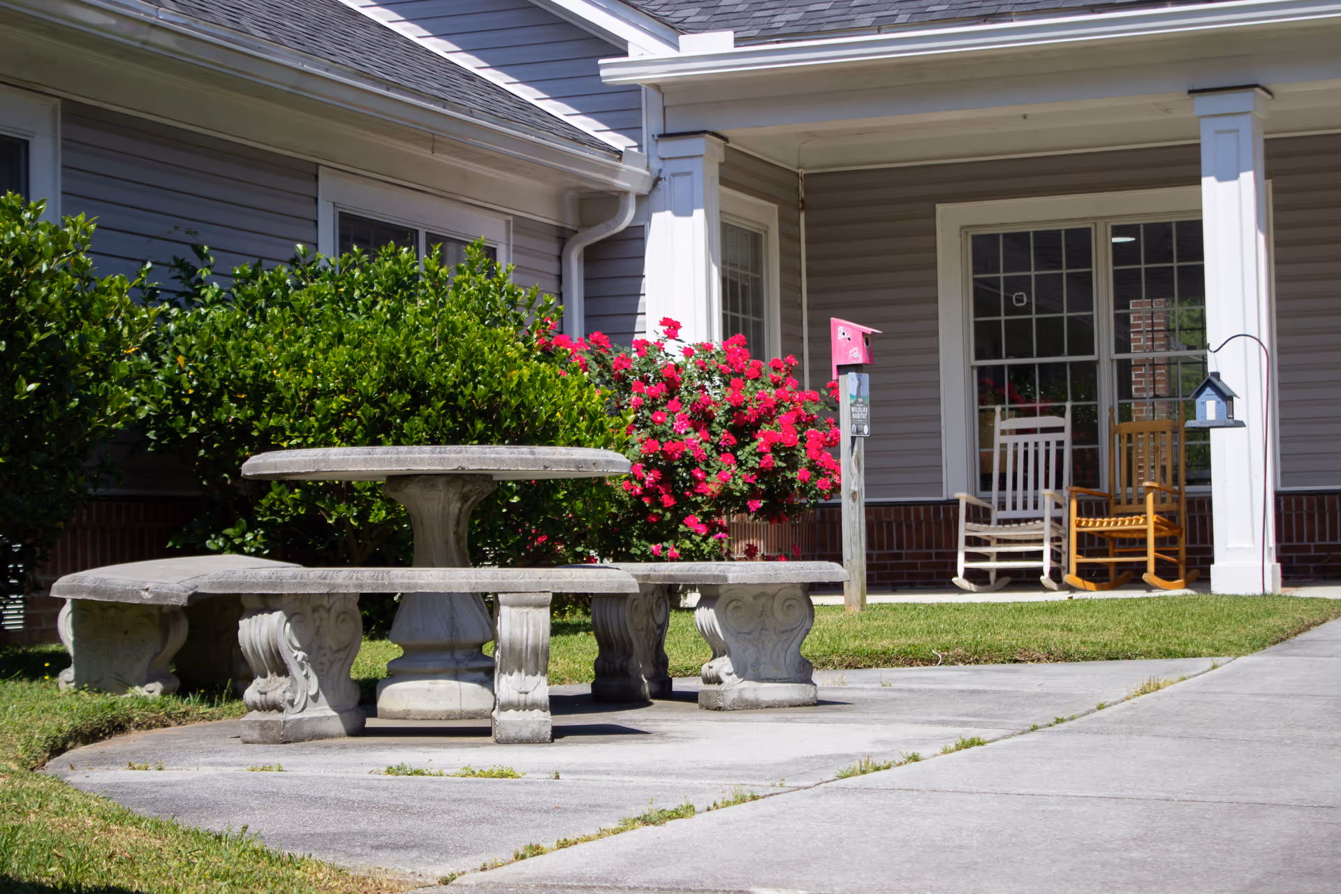 Stone picnic table and benches on a concrete patio in front of a building porch with rocking chairs and flowering bushes.