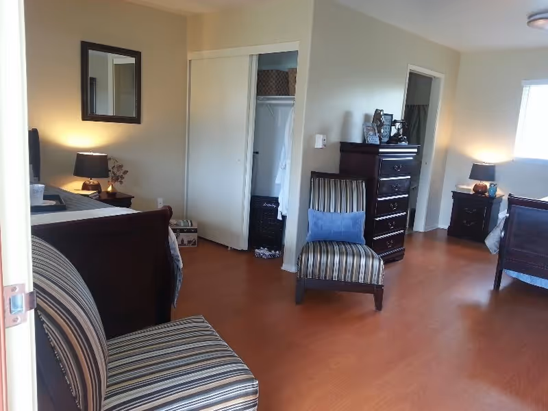 A spacious bedroom with wooden flooring featuring two striped armchairs, two dark wooden beds with white bedding, two nightstands with lamps, a tall dresser, an open closet with hanging robes, and a window letting in natural light.