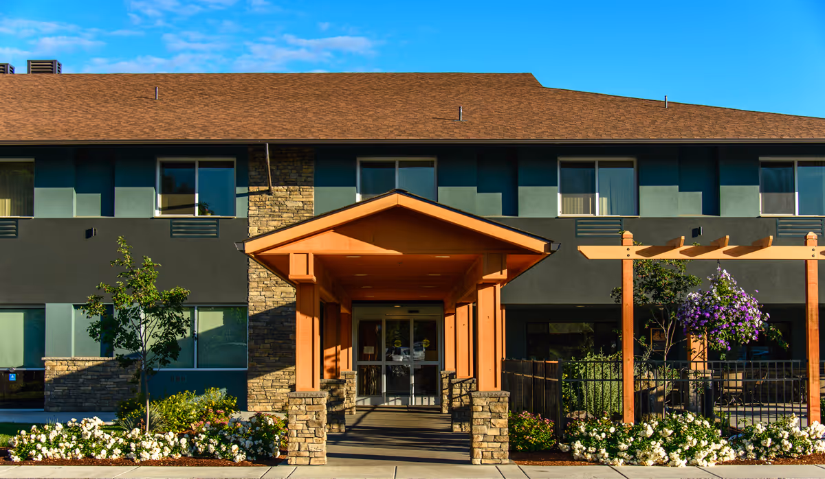 Front exterior view of a two-story building with a brown roof, green and gray walls, and a covered entrance supported by stone pillars. There are windows on both floors and landscaped flower beds with white and purple flowers along the walkway leading to the entrance.