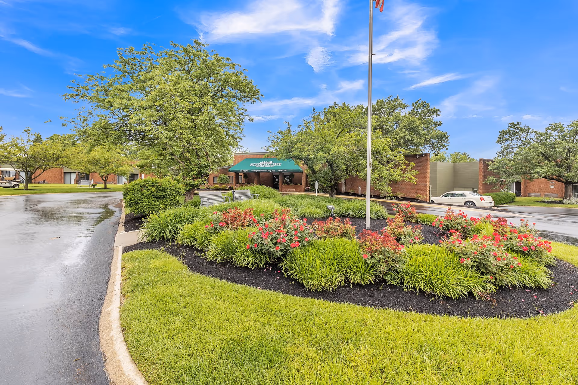 View of the entrance to Northgate Park facility with a circular flower bed featuring red flowers and green shrubs in the foreground, a flagpole in the center, and a driveway curving around the landscaped area. The building is a single-story brick structure with a green awning over the entrance. Trees and a partly cloudy blue sky are visible in the background.