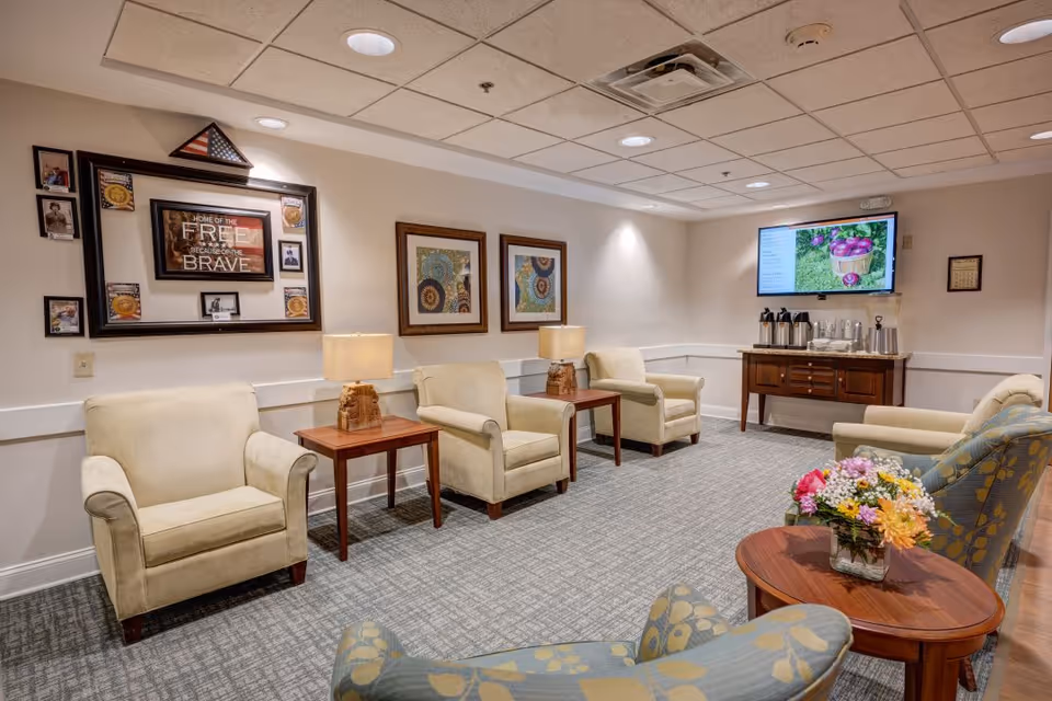 A cozy sitting area with beige armchairs and wooden side tables, decorated with framed artwork and a patriotic display on the wall. A flat-screen TV is mounted on the far wall above a wooden cabinet with coffee and water dispensers. A small round table with a vase of colorful flowers is in the foreground.