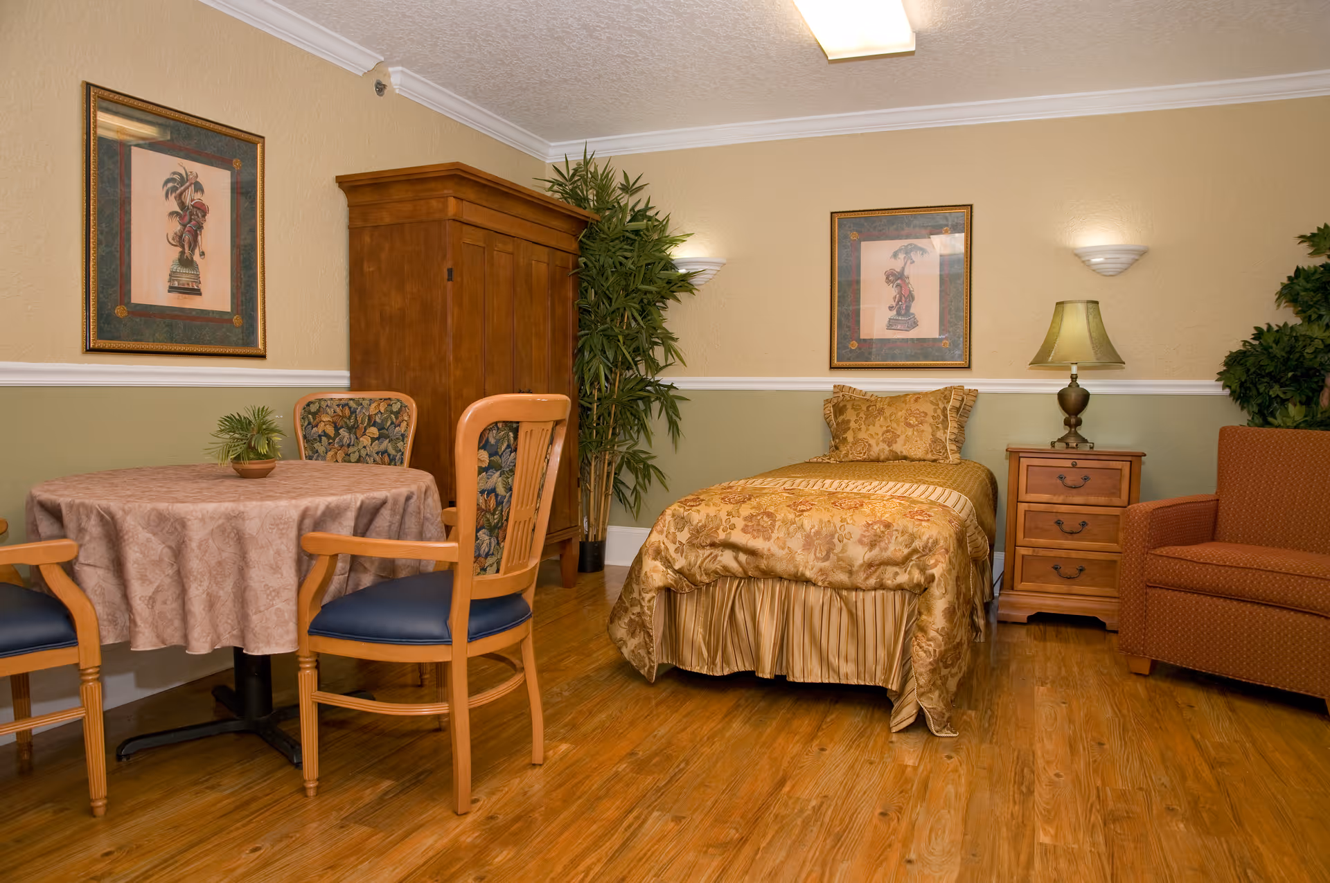 A cozy room with a single bed covered in a gold and floral patterned bedspread, a wooden nightstand with a lamp, an orange armchair, a wooden armoire, a round table with a beige tablecloth, and three wooden chairs with floral cushions. The walls are painted beige and green with two framed pictures and two wall-mounted lights. There are also two potted plants in the corners.
