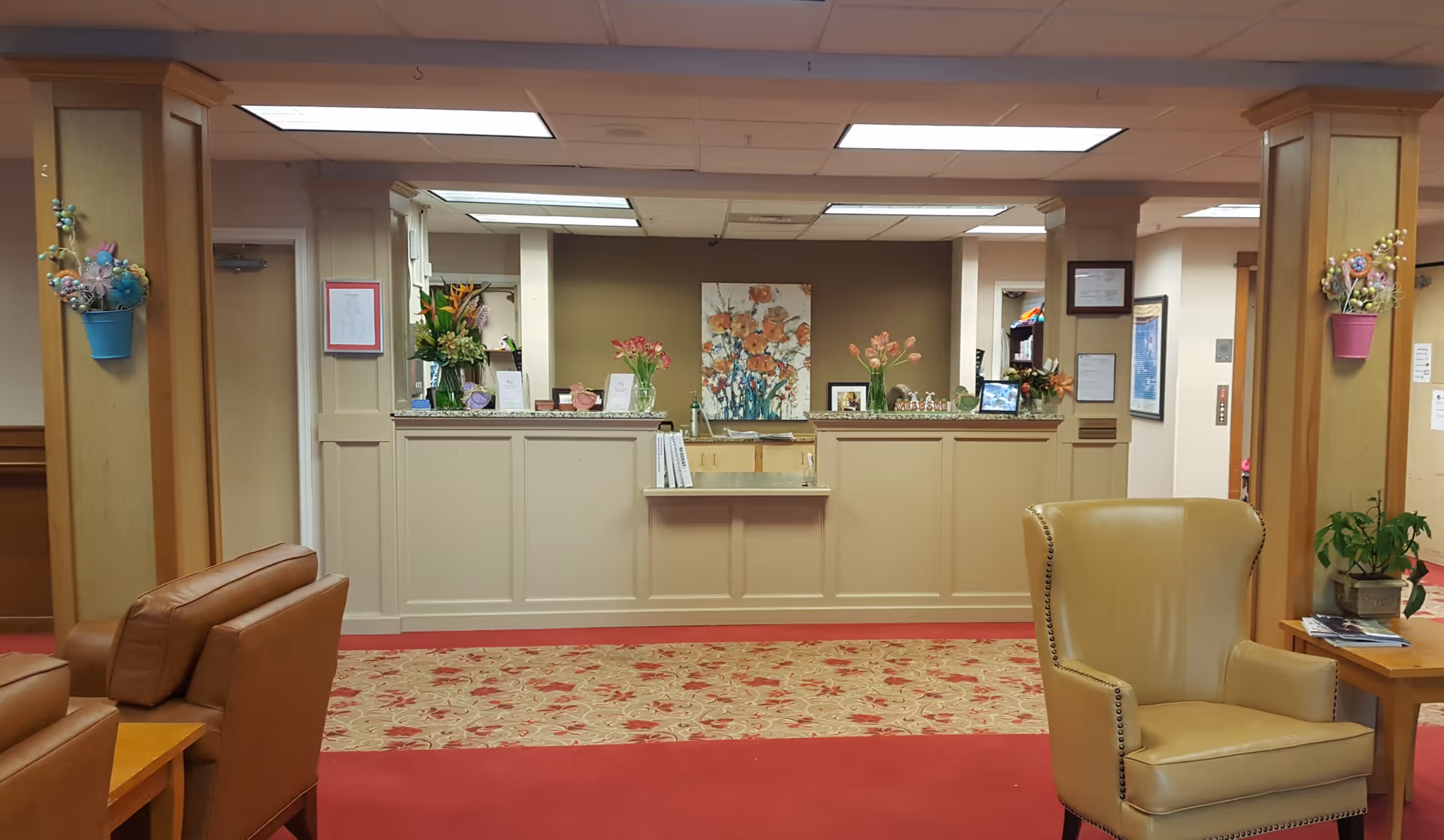 Reception area of an assisted living facility with a front desk decorated with flower vases and a floral painting on the wall behind. The room has a red carpet with a floral pattern, leather chairs, wooden pillars with colorful flower decorations, and a small table with a plant and magazines.
