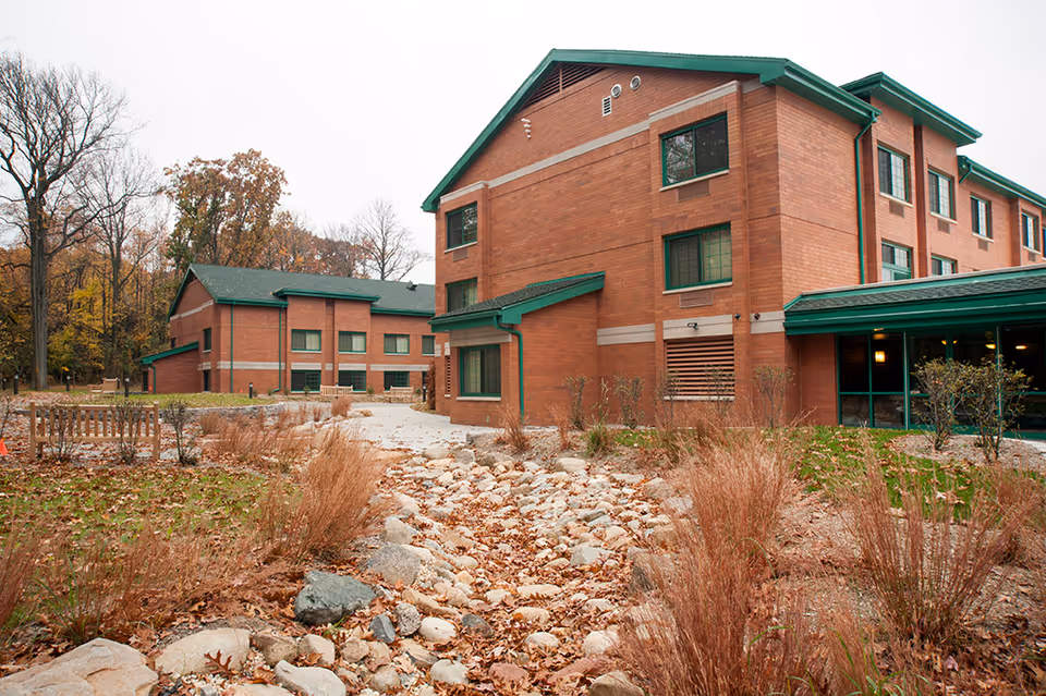 Exterior view of a brick multi-story senior living building with green trim beside a landscaped dry rock bed and fall foliage.