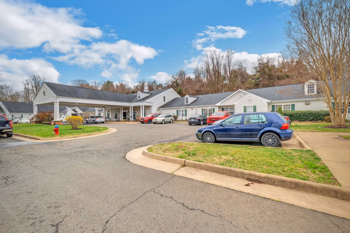 Exterior view of a senior living facility with a covered entrance, multiple parked cars, and a surrounding landscape with trees and grass under a partly cloudy sky.