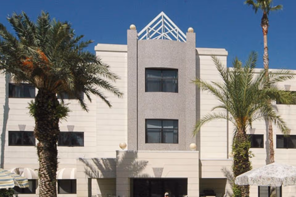 Exterior view of a multi-story building with a modern design, featuring palm trees in front and a clear blue sky in the background.