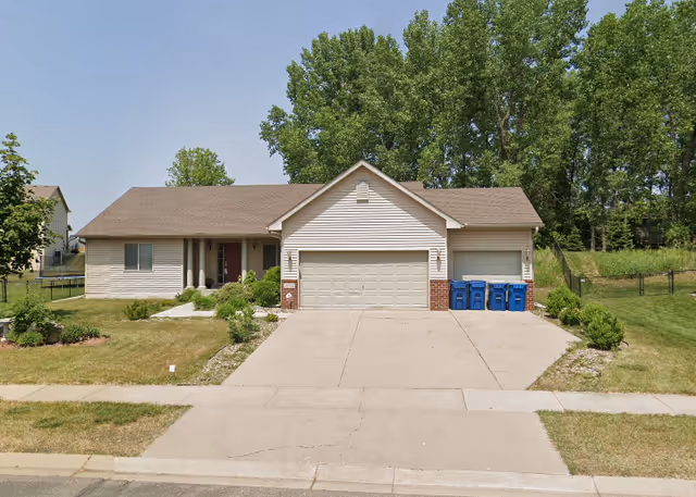 Front exterior view of a single-story house with a three-car garage, a concrete driveway, and a well-maintained lawn with shrubs and trees in the background under a clear sky.