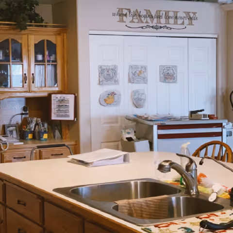 Interior view of a kitchen area in an assisted living facility featuring a double sink with a faucet on a countertop island. Behind the island, there is a wooden cabinet with glass doors and various items on the counter. On the wall, there are white closet doors decorated with children's drawings and a wall decal above that reads 'FAMILY where life begins and love never ends.'