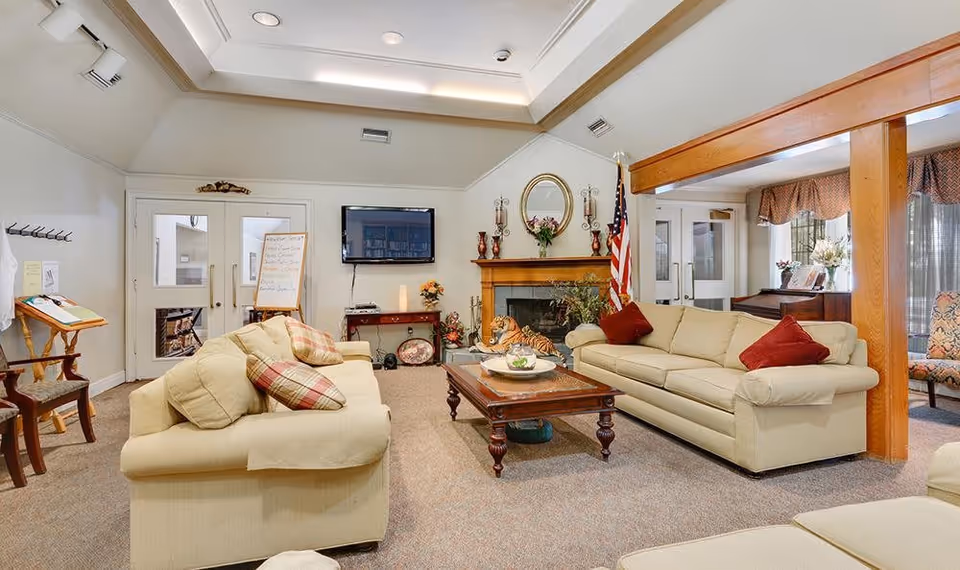 A cozy living room in a senior living facility with two beige sofas adorned with red and plaid pillows, a wooden coffee table, a fireplace with decorative items and an American flag beside it, a wall-mounted TV, and a piano near a window with curtains.