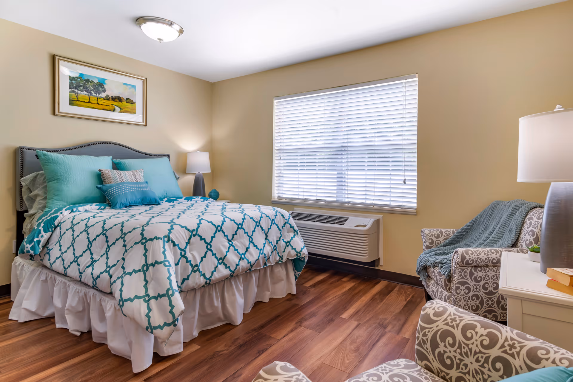 A neatly furnished bedroom with a bed dressed in teal-accented bedding, two armchairs, bedside lamps, and a window with blinds.