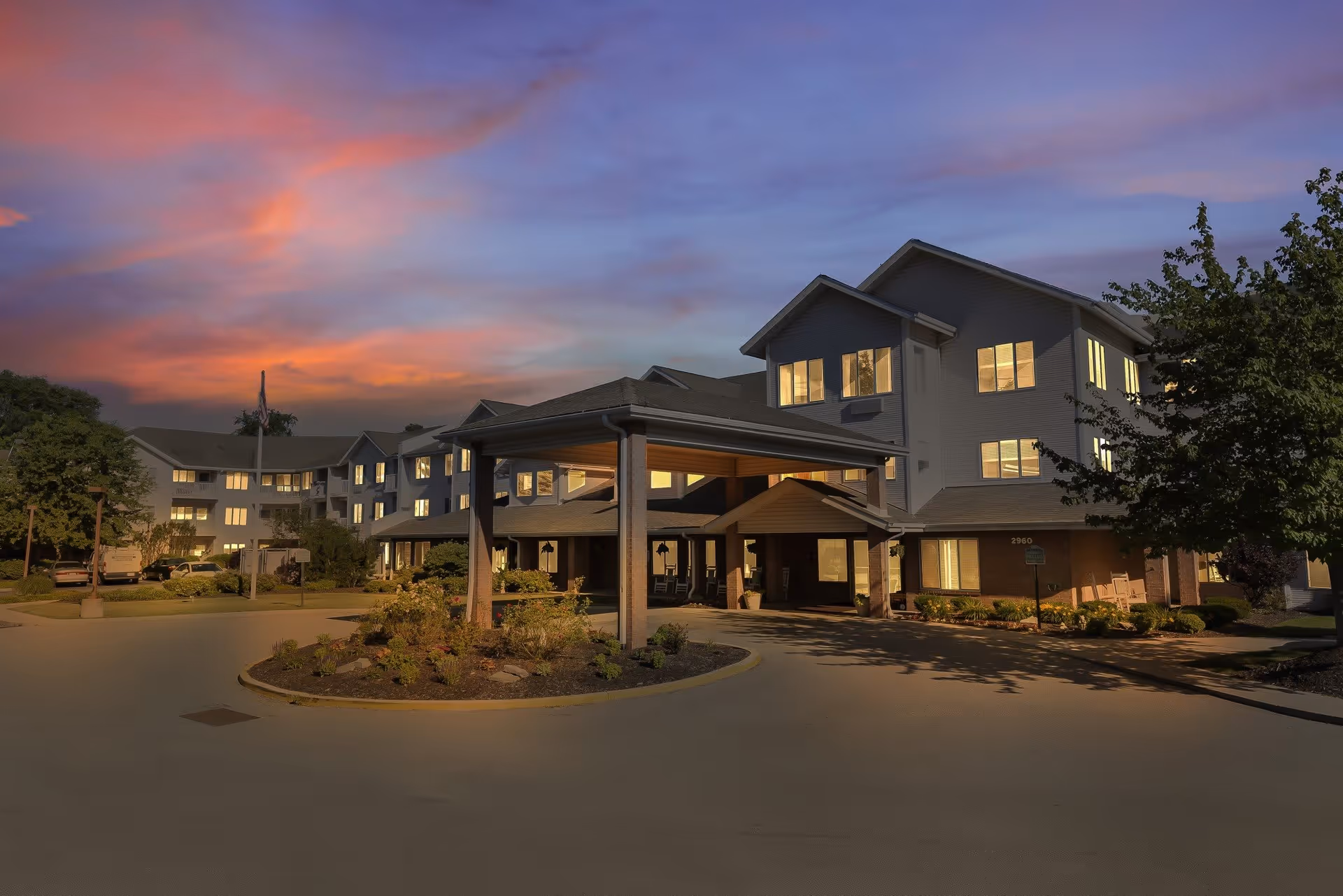 Exterior view of a multi-story senior living facility named Provincial Bethel Park at dusk, with warm lights glowing from the windows and a covered entrance surrounded by landscaped greenery.