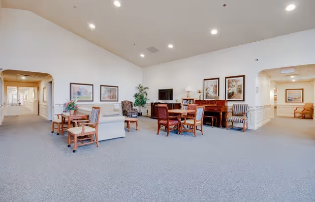 A spacious, well-lit common area in a senior living facility with multiple seating arrangements including armchairs and small tables. The room features a piano against the wall, framed artwork, a television, and decorative plants. Two hallways are visible on either side of the room.