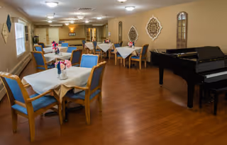 A spacious room with several tables covered with white tablecloths and blue cushioned chairs arranged around them. Each table has a small floral centerpiece. The room has wooden flooring, beige walls with decorative wall hangings, and a black grand piano on the right side. Ceiling lights illuminate the area.