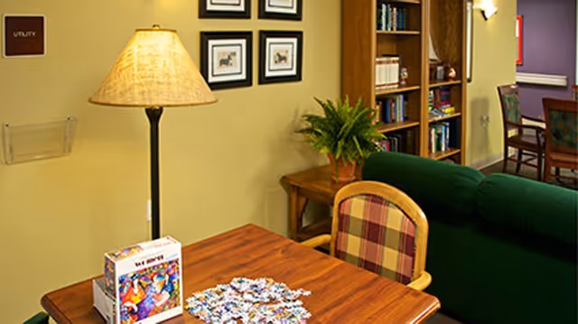 Cozy seating area with a wooden table holding a jigsaw puzzle and box, a floor lamp, upholstered chairs, a green sofa, and bookshelves.