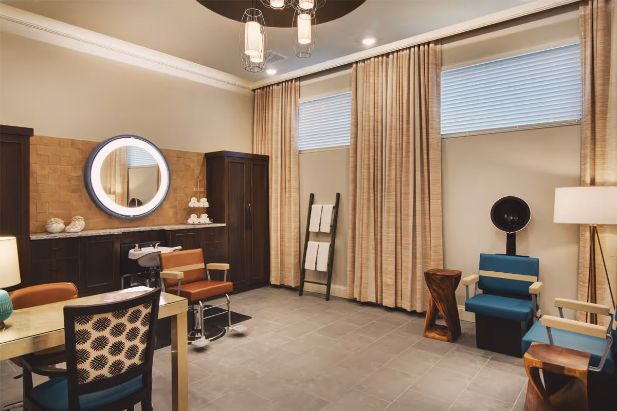 Interior view of a stylish salon room with a hair washing station, two salon chairs, a round illuminated mirror, a wooden cabinet, a towel rack with white towels, and two blue chairs with a hair dryer hood. The room has beige curtains, tiled floor, and modern lighting fixtures.