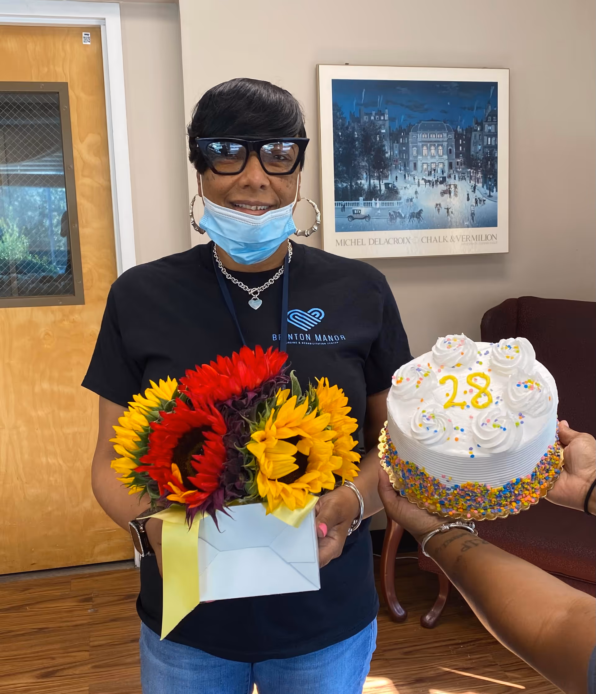 A woman wearing glasses, a face mask under her chin, and a black Brinton Manor Nursing and Rehabilitation Center t-shirt is holding a bouquet of sunflowers and red flowers in a white box with a yellow ribbon. Another person is holding a white frosted cake decorated with colorful sprinkles and the number 28 written in yellow icing. They are indoors with a wooden door and a framed painting in the background.