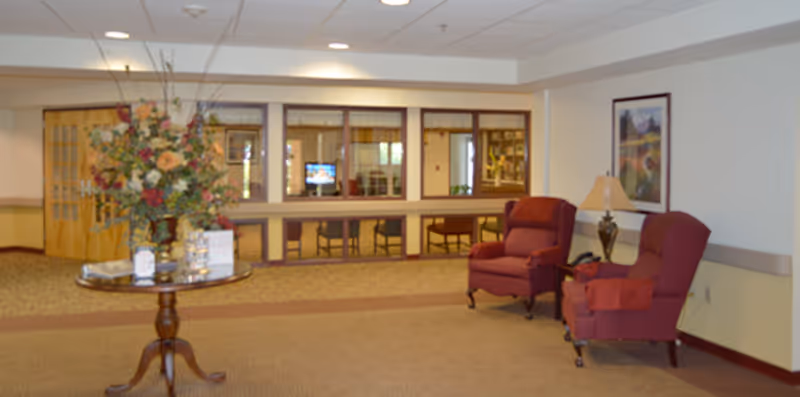 A spacious common area in an assisted living center featuring a round wooden table with a large floral arrangement in the center. Two upholstered armchairs with a small side table and lamp are positioned against the wall. Large windows look into an adjacent room with chairs and a television visible.