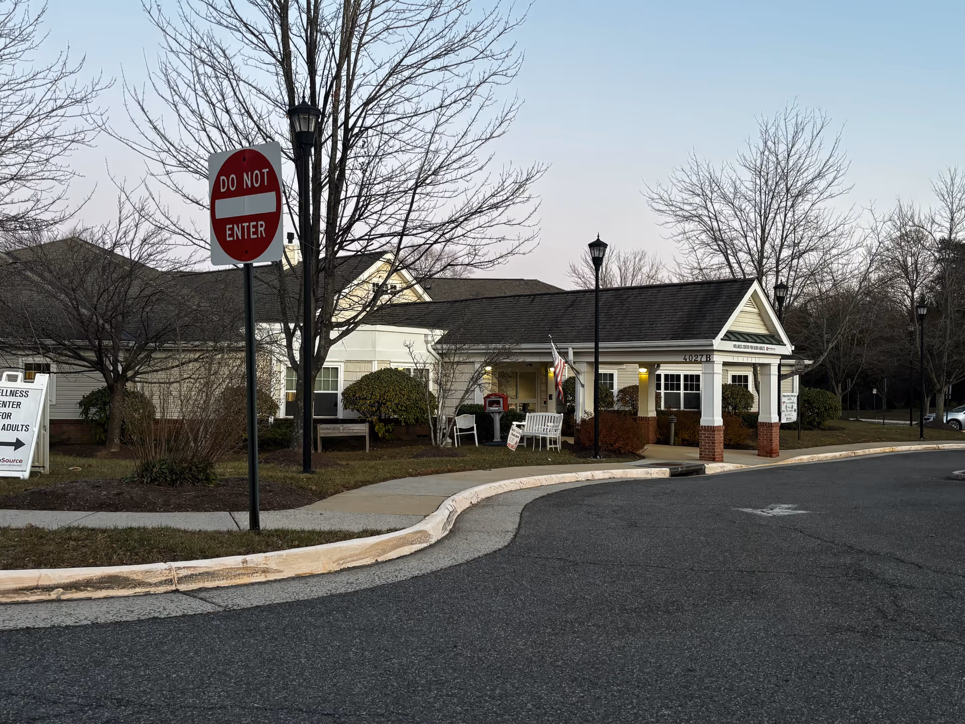 Exterior view of a single-story building with a covered entrance, white columns, and brick accents. There are leafless trees and bushes around the building, a 'Do Not Enter' sign on a pole, and a sidewalk curving around the front. The sky is clear and it appears to be either early morning or late afternoon.