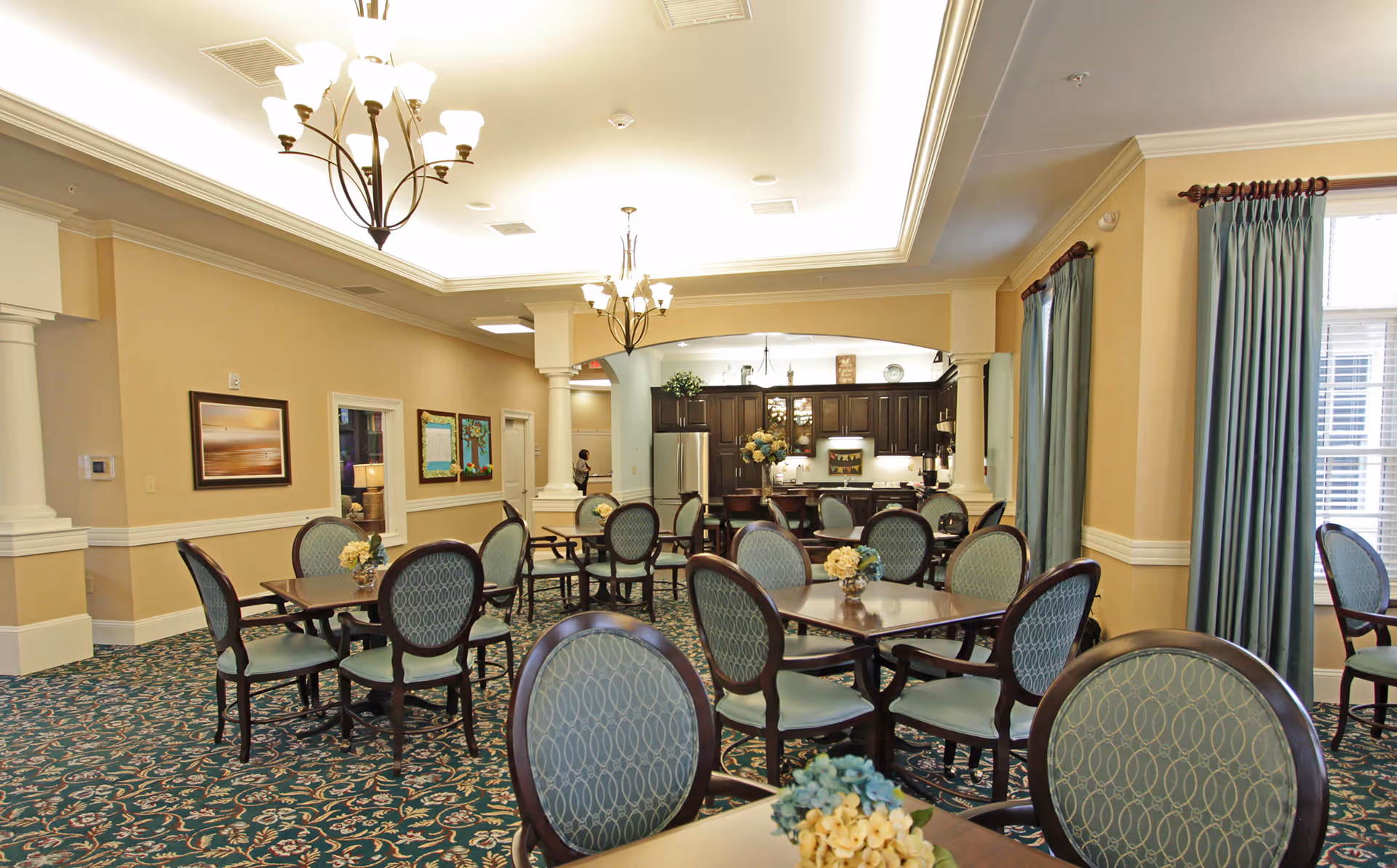 A well-lit dining room with multiple tables and chairs arranged neatly. The chairs have blue patterned upholstery and dark wooden frames. The room features a patterned carpet, beige walls, and large windows with blue curtains. In the background, there is a kitchen area with dark wooden cabinets and stainless steel appliances. Two chandeliers hang from the ceiling providing warm lighting.