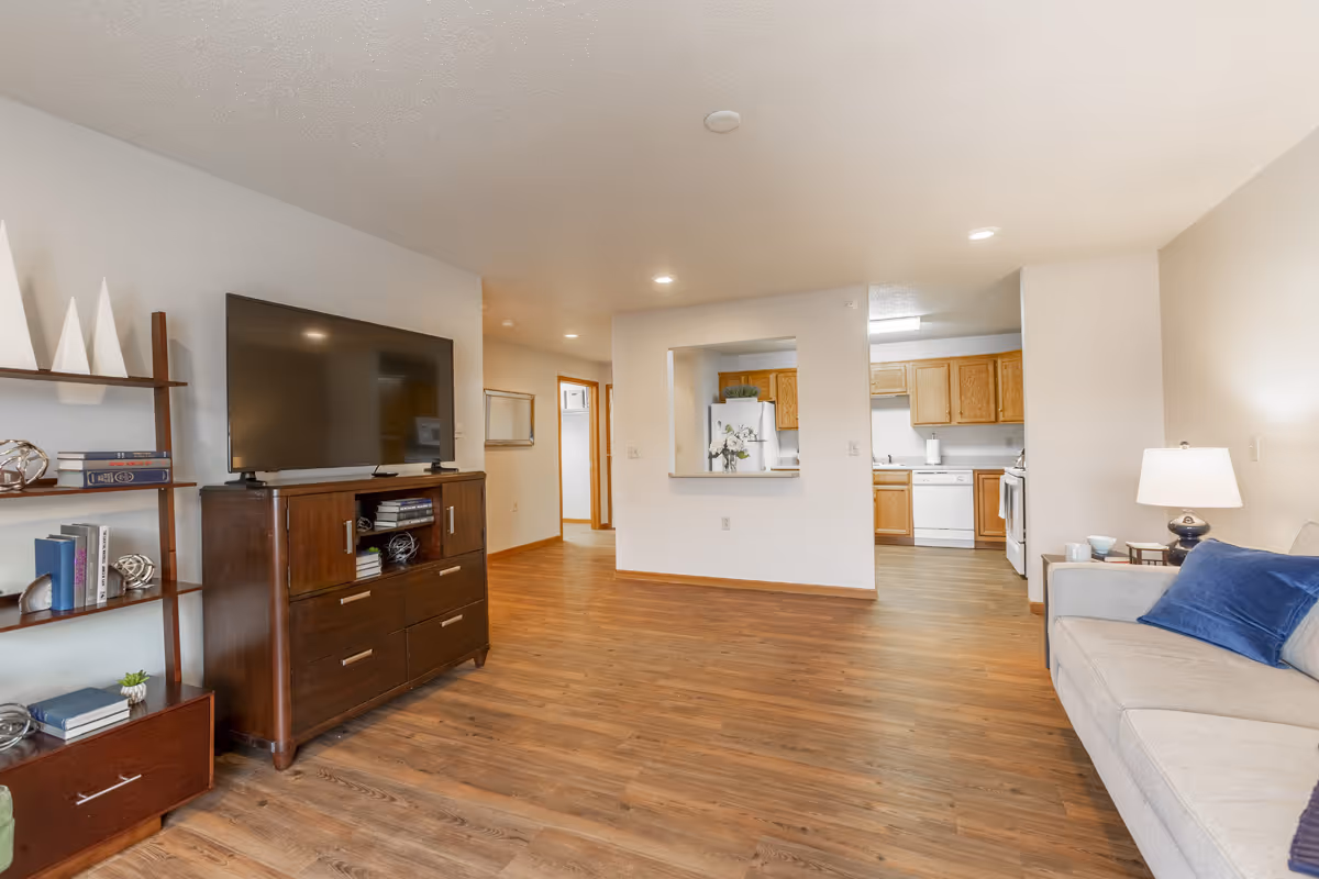 A spacious living room with wooden flooring featuring a large flat-screen TV on a dark wooden cabinet, a bookshelf with decorative items and books, a beige sofa with blue cushions, and a lamp on a side table. The room opens into a kitchen area with wooden cabinets, a white refrigerator, dishwasher, and stove.
