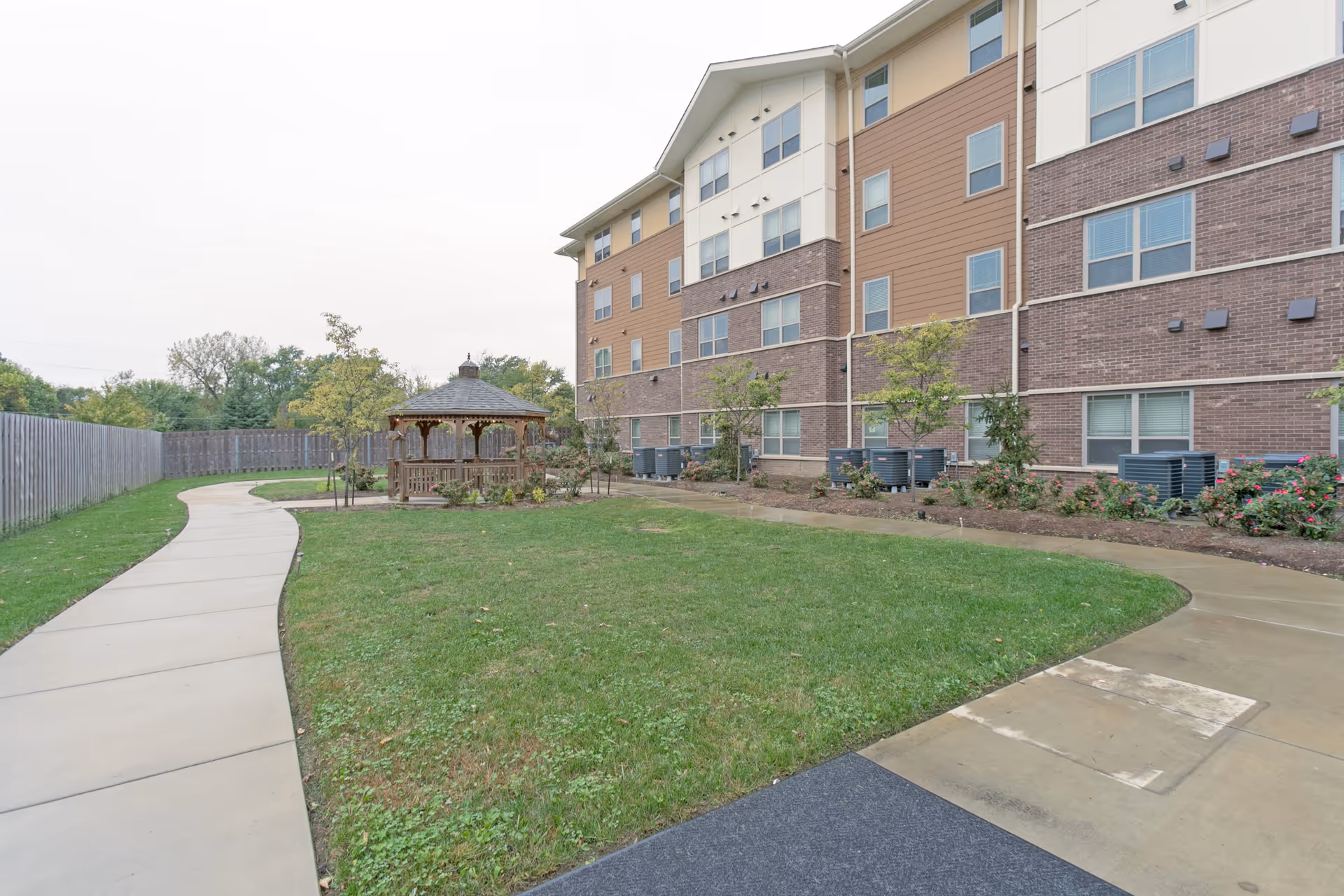 Outdoor area of Oasis At 30th featuring a curved concrete walkway, a grassy lawn, a wooden gazebo, and a multi-story building with brick and siding exterior in the background.
