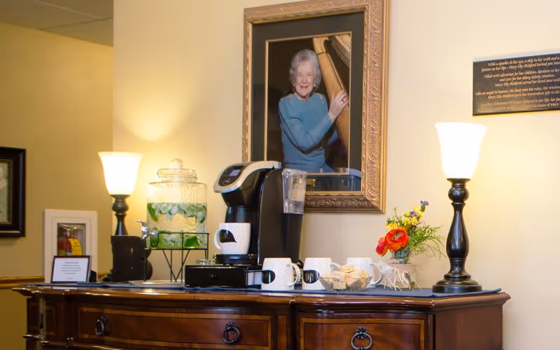 A wooden sideboard with a coffee maker, several white mugs, a glass bowl with crackers, a glass dispenser with water and cucumber slices, two table lamps with white shades, a small flower arrangement, and a framed portrait of an elderly woman playing a harp hanging on the wall behind.