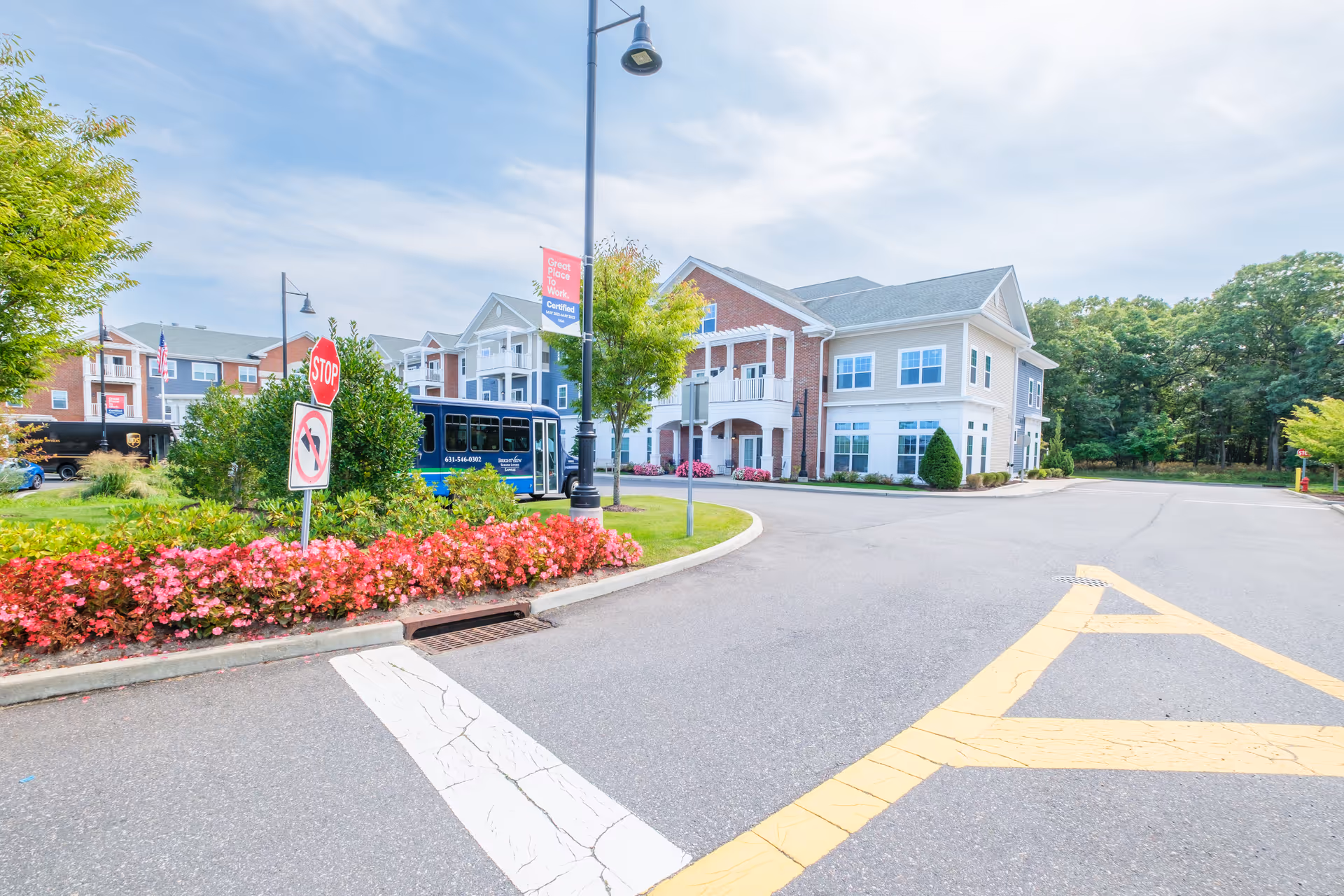 Exterior view of a senior living facility with a paved driveway, landscaped flower beds with pink flowers, a stop sign, a blue shuttle bus, and a multi-story building with balconies and large windows under a partly cloudy sky.