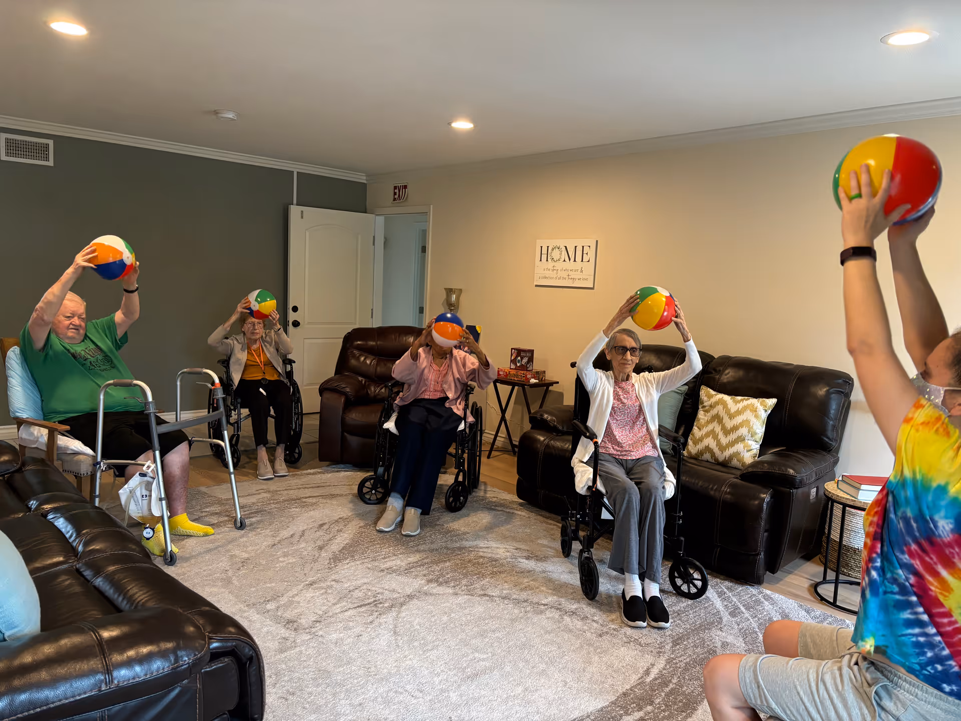 A group of elderly individuals seated in a living room area participating in a seated exercise activity, each holding a colorful beach ball above their heads. The room has leather sofas, a patterned rug, and a wall sign that reads 'HOME'.