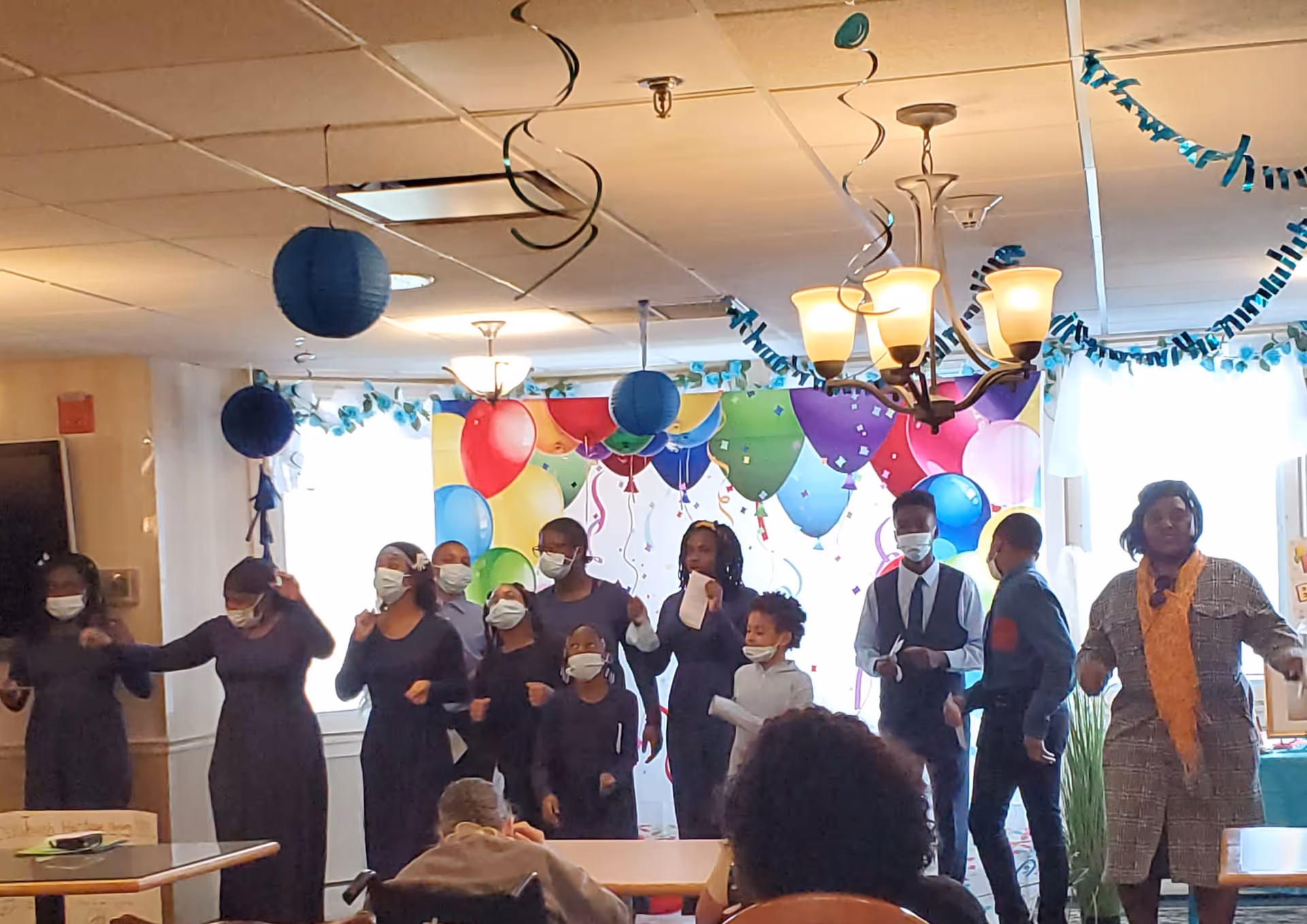 A group of people, including children and adults wearing masks, standing and performing in a decorated room with colorful balloon-themed wall art and hanging blue paper decorations. Some audience members are seated and watching the performance.