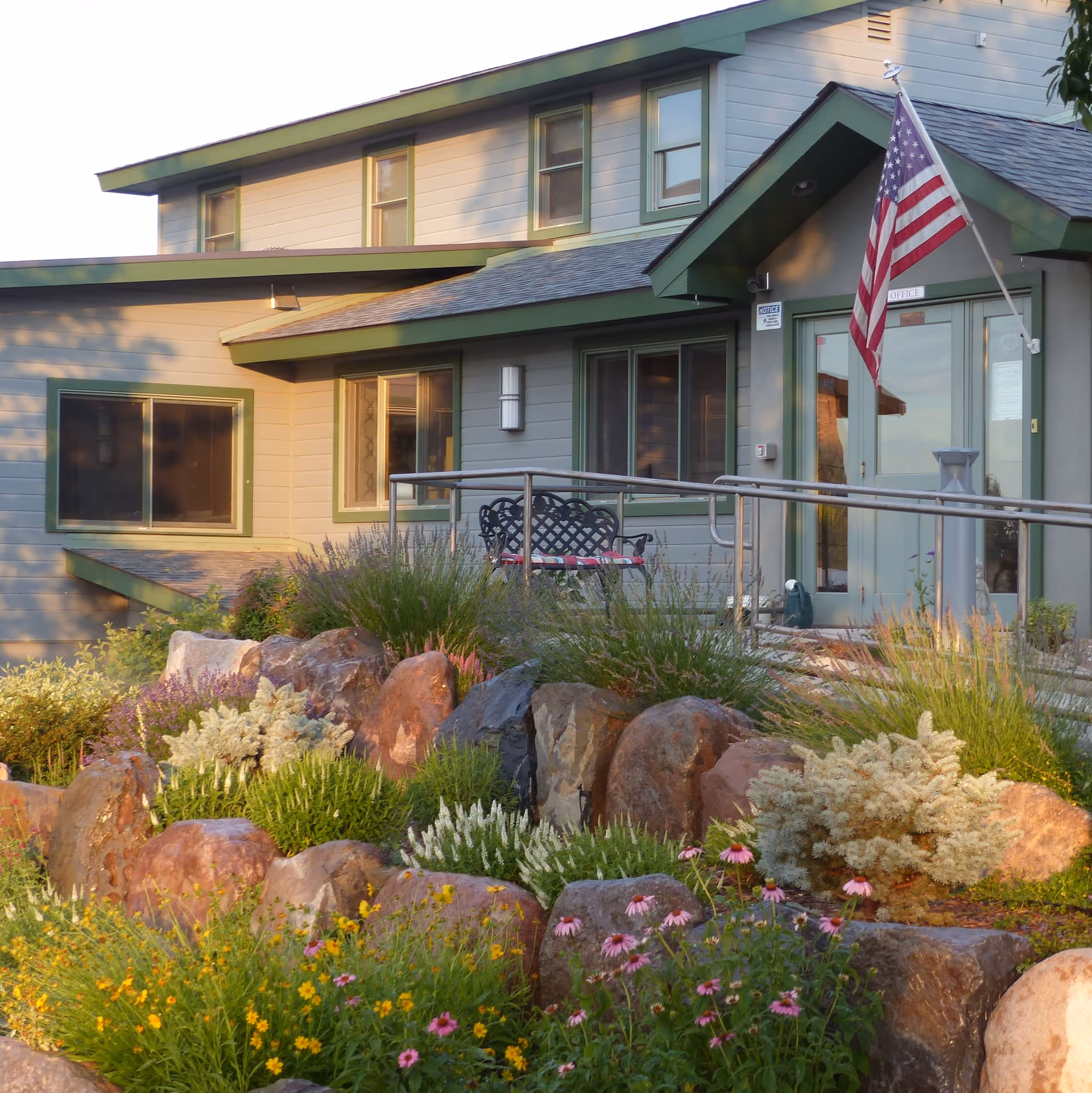 Exterior view of Evenings Porch Assisted Living facility showing a building with green-trimmed windows and doors, an American flag near the entrance, a ramp with metal railings, and a landscaped garden with rocks and various flowering plants in the foreground.