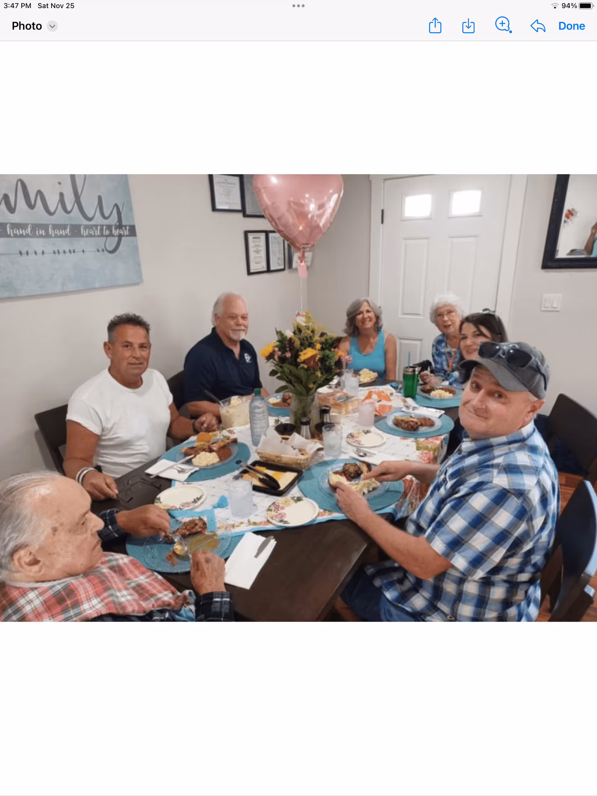 A group of six adults sitting around a dining table enjoying a meal together. The table is set with plates of food, drinks, and a centerpiece of flowers with a pink heart-shaped balloon. The room has light-colored walls with framed pictures and a door in the background.