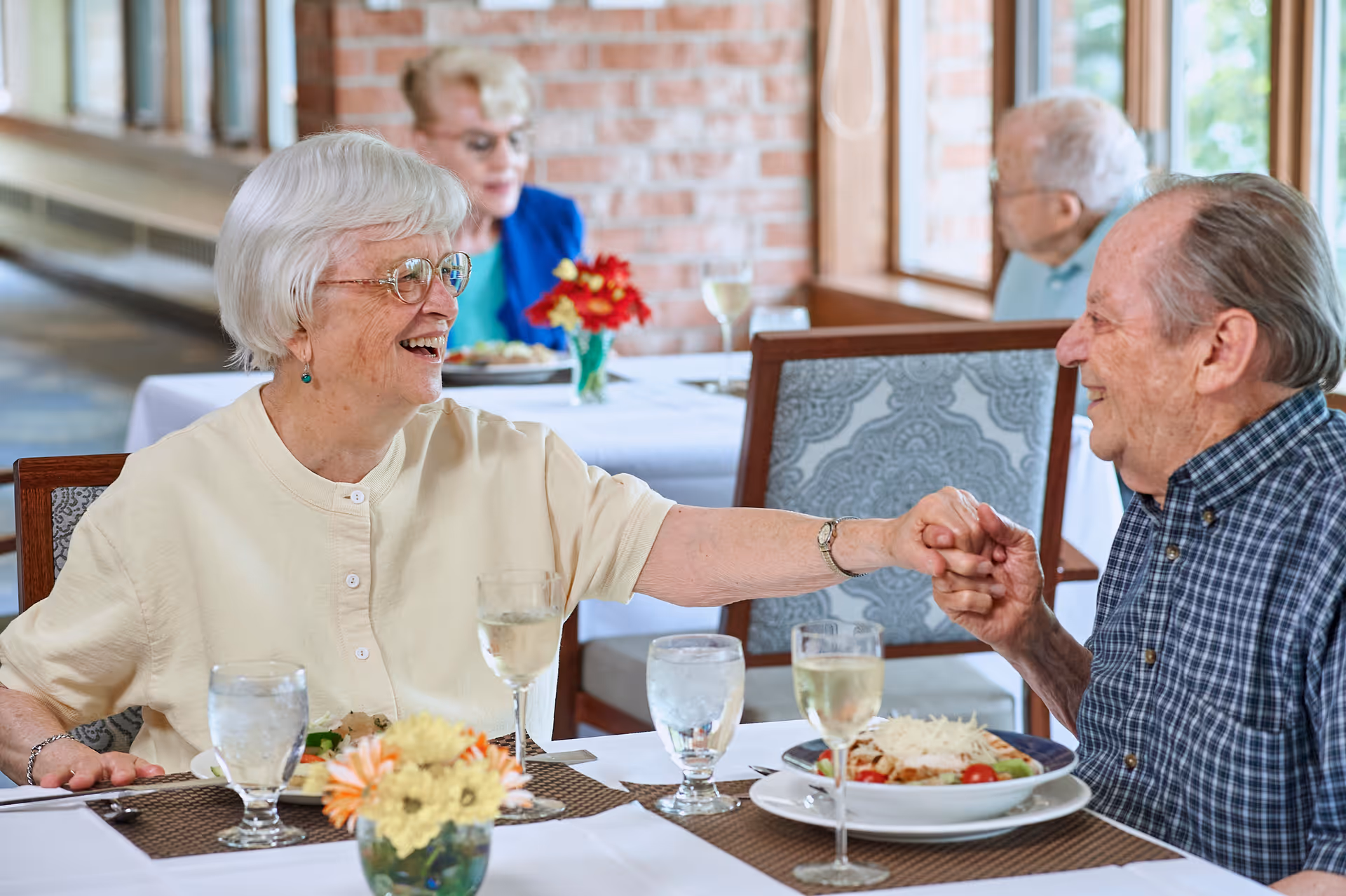 An elderly woman and man smile and hold hands across a dining table with plates and glasses in a senior living dining room.