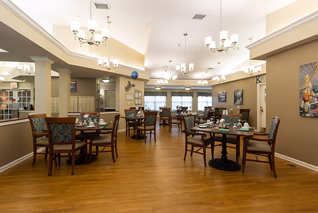 A spacious dining room with multiple wooden tables and chairs arranged neatly on a wooden floor. The tables are set with cups, saucers, and small flower arrangements. The room is well-lit with multiple chandeliers hanging from the ceiling. Large windows with curtains allow natural light to enter. The walls are decorated with framed pictures and artwork.