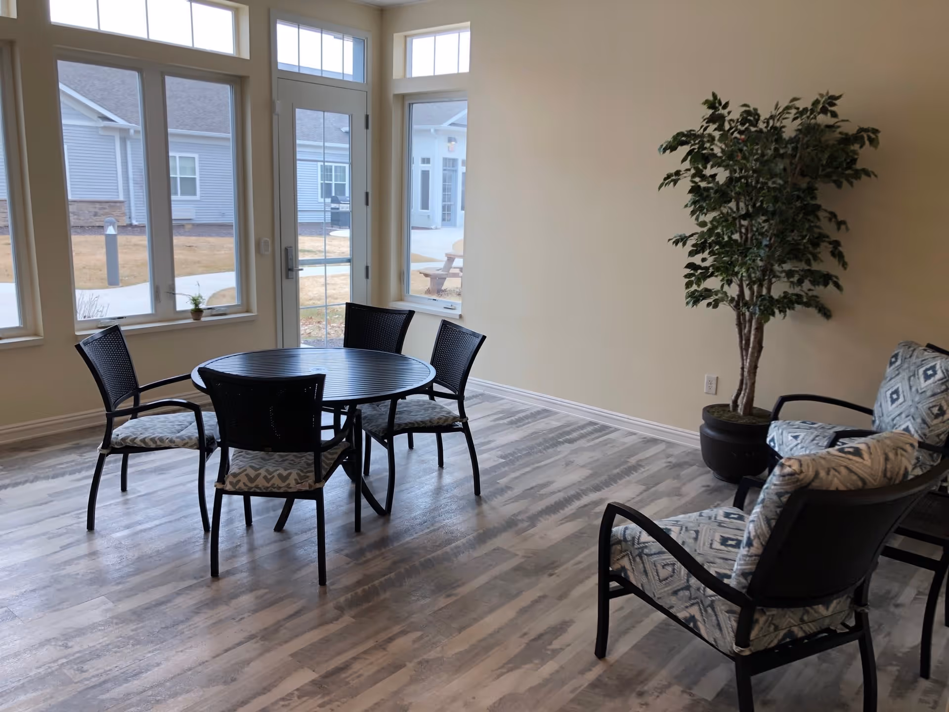 A bright room with large windows and a glass door leading outside. Inside, there is a round black table with four black chairs around it, each with patterned cushions. To the right, there are two cushioned armchairs with a geometric pattern and a tall potted plant next to them. The floor has a light wood pattern and the walls are painted beige.