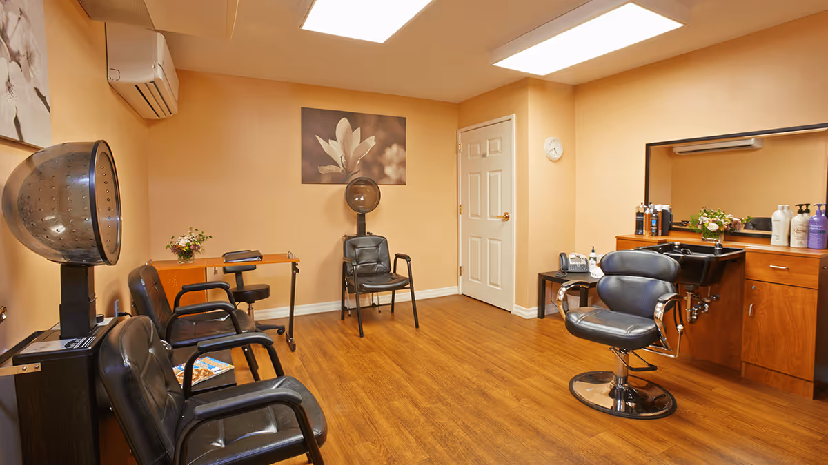 Interior of a salon room with wooden flooring, beige walls, and ceiling lights. The room contains black salon chairs, a hair dryer, a sink with hair care products on the counter, a small table with a flower vase, and a wall clock above a closed door. A large mirror is mounted above the sink area.