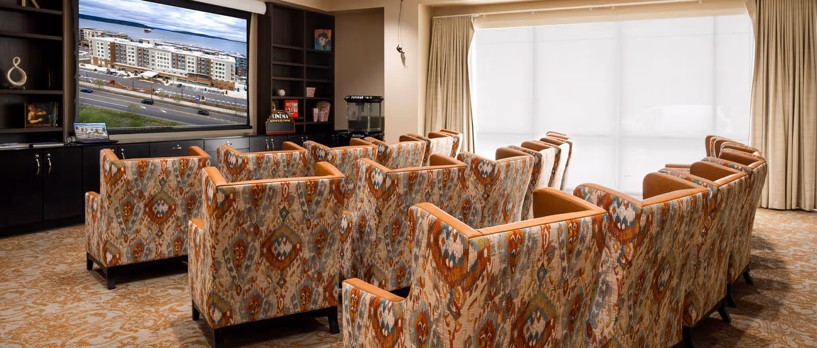 A cozy media room with multiple patterned armchairs arranged in rows facing a large flat-screen TV mounted on a dark built-in cabinet. The room has beige carpet and large windows covered with light curtains, allowing natural light to softly illuminate the space.