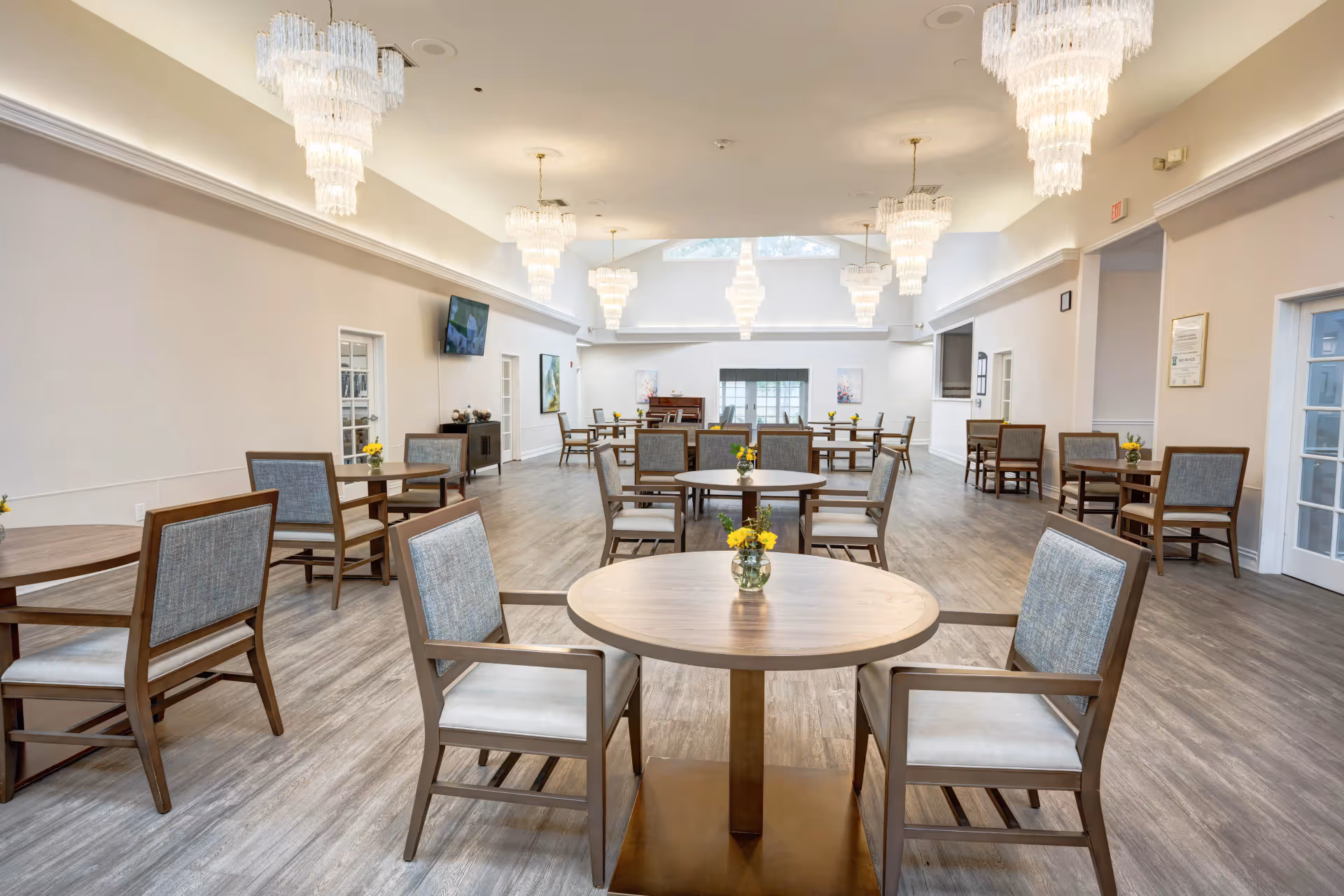 A spacious dining room with multiple round wooden tables, each surrounded by four cushioned chairs. The room features elegant chandeliers hanging from the ceiling, light-colored walls, and large windows at the far end allowing natural light to enter. Small flower arrangements are placed on each table, and a television is mounted on one wall.